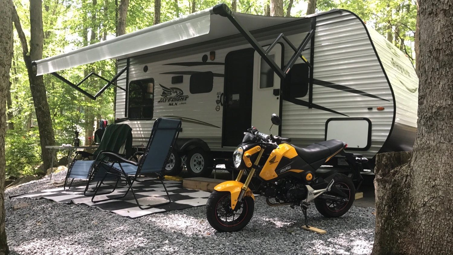 Exterior of Jay Flight with Chairs and a motorcycle in front of it. In a wooded campsite.