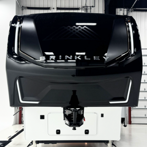 Front cap of a Brinkley fifth wheel RV inside a service bay, showcasing a sleek, glossy black finish with integrated LED light accents, bold BRINKLEY branding, and a modern aerodynamic design.