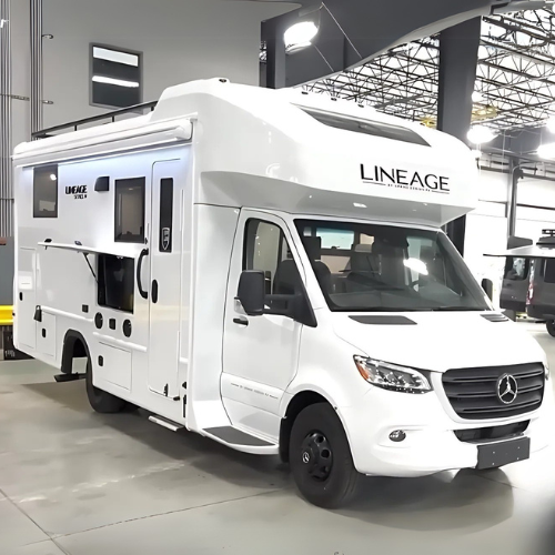 Front-side view of a Grand Design Lineage 25TK Class C motorhome inside a manufacturing facility, built on a white Mercedes-Benz Sprinter chassis with LINEAGE branding above the cab, featuring exterior storage compartments, an outdoor TV, and a sleek, modern design.