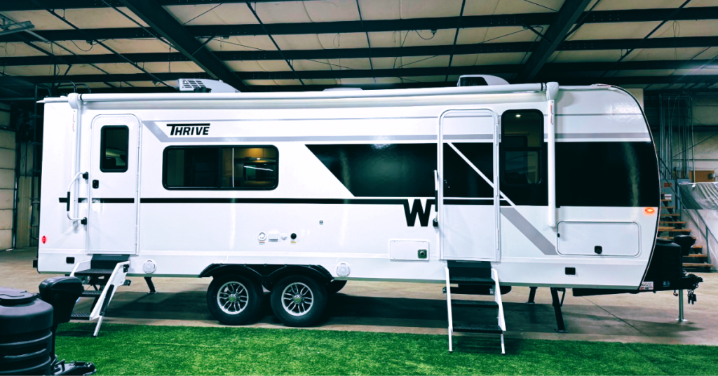 Side view of a Winnebago Thrive travel trailer displayed indoors, featuring a bold black-and-white exterior design, dual axles, two entry doors, and retractable steps on artificial turf.
