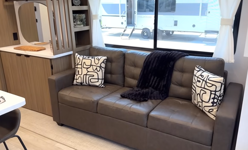Stylish RV living area featuring a tufted dark brown leather sofa with modern abstract print pillows and a soft black throw blanket, set beneath a large window with white curtains, adjacent to wood cabinetry and built-in shelving.