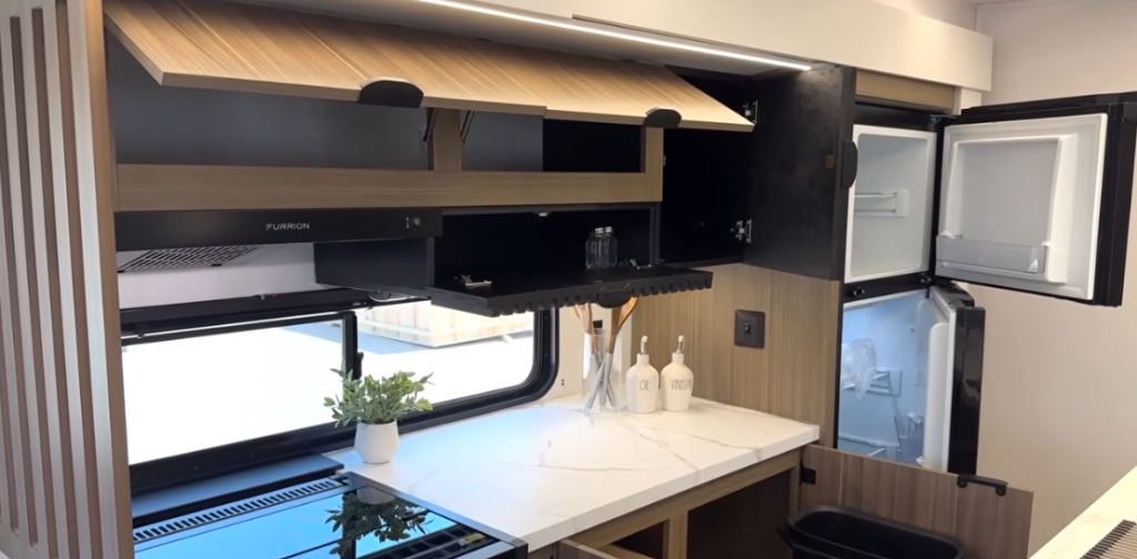 RV kitchen area with upper cabinets open to reveal glassware and shelving, a white marble-style countertop with a potted plant and oil and vinegar containers, an open two-door refrigerator, a black stovetop, and a hidden pull-out trash bin beneath the counter.
