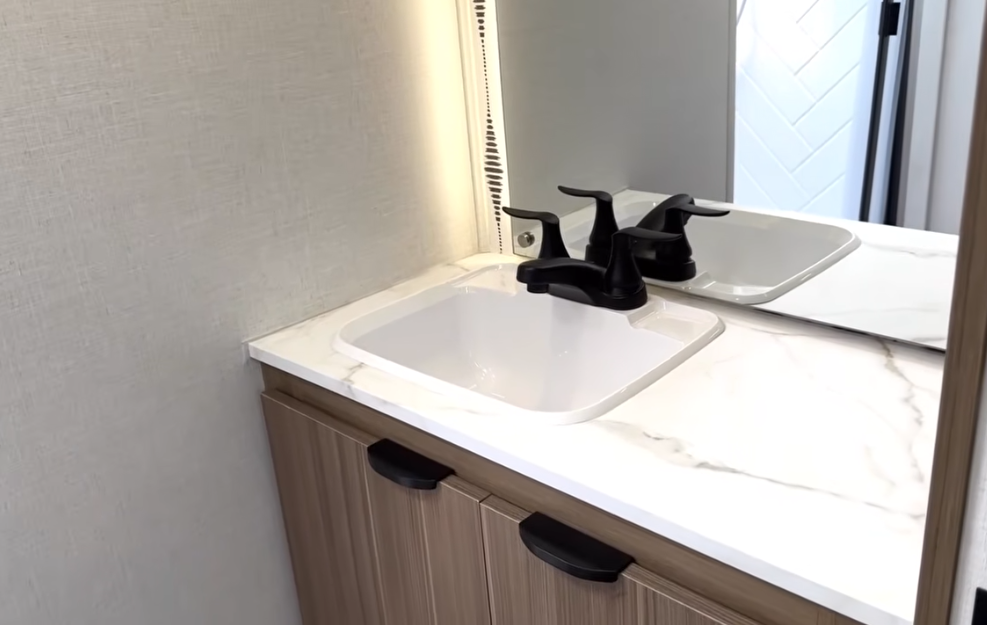 Split-screen image showing a man in a Bish's RV polo shirt giving a thumbs-up on the left side inside an RV shower, with the right side displaying a bright, curved RV shower stall featuring white herringbone tile walls, black trim, a skylight, and a handheld showerhead.