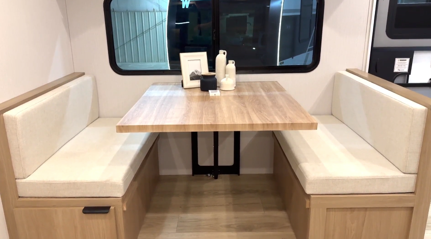 A modern RV dinette with light wood bench seating and beige cushions, featuring a center-mounted wooden table set in front of a large window. Simple decorative items sit on the table.