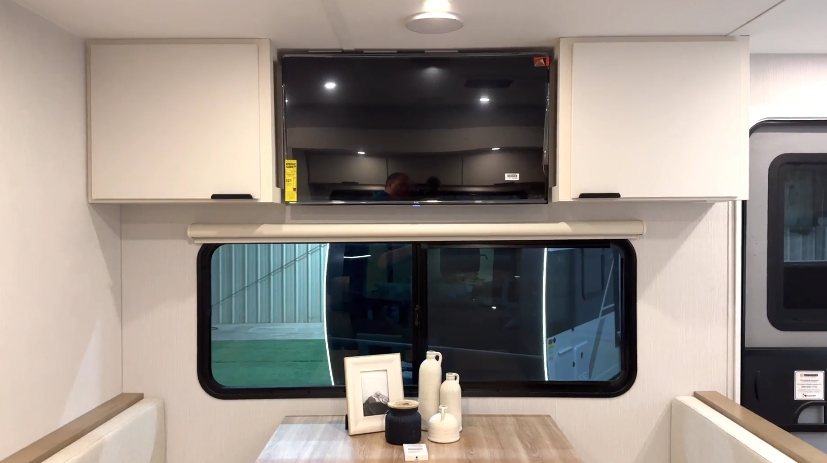 Interior view of an RV showing a mounted TV above a large window with closed blinds, positioned above a dinette table with decorative items. Light cabinetry surrounds the TV.