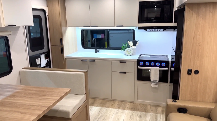 Modern RV kitchen interior with a large window over a countertop, black oven with glowing blue knobs, built-in microwave, upper and lower cabinets, a booth dinette with a wooden table, and part of a tan recliner.