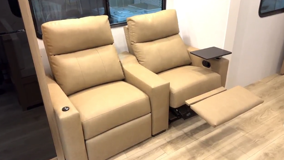  A pair of tan RV recliners with plush cushions and one extended footrest, featuring a swivel tray table and cup holder, positioned by a window inside a travel trailer.