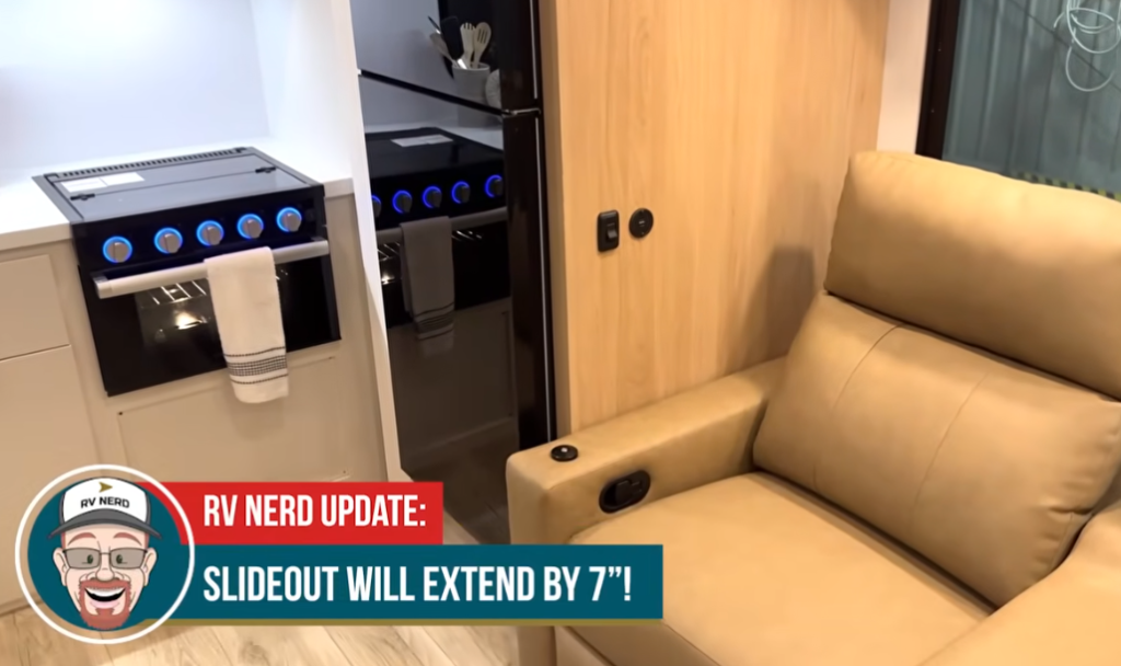 Interior of an RV showing a tan recliner next to a wood-paneled wall and a kitchen area with a black oven, blue-lit knobs, and a refrigerator; text overlay notes a 7" slideout extension update.