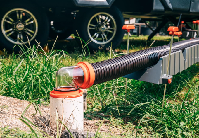 An RV sewer hose connected to a campground dump station. The black, accordion-style hose is supported by a sloped plastic hose support system with orange caps, leading from the RV to a white PVC sewer inlet. A clear elbow fitting with orange connectors allows visibility of waste flow, ensuring a secure and clean connection for dumping the RV’s holding tanks. Grass and RV tires are visible in the background.