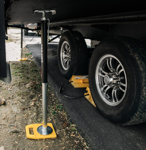 An RV stabilization setup showing two trailer tires with yellow wheel chocks and a telescoping steel stabilizer jack placed between the axles. The jack rests on a yellow stabilizer pad for added support and stability. The scene is set on a paved and dirt surface, with the underside of the RV visible above the equipment.
