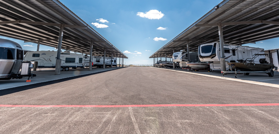 A row of RVs and trailers parked under large covered storage structures on both sides of a wide asphalt lot, with a clear blue sky and scattered clouds in the background. The image depicts a secure and spacious covered RV storage facility.