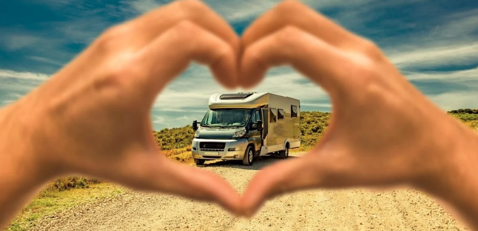 A motorhome is seen through a heart shape made with two hands in the foreground, symbolizing a love for RV travel. The RV is parked on a dirt road surrounded by open landscape and blue skies.