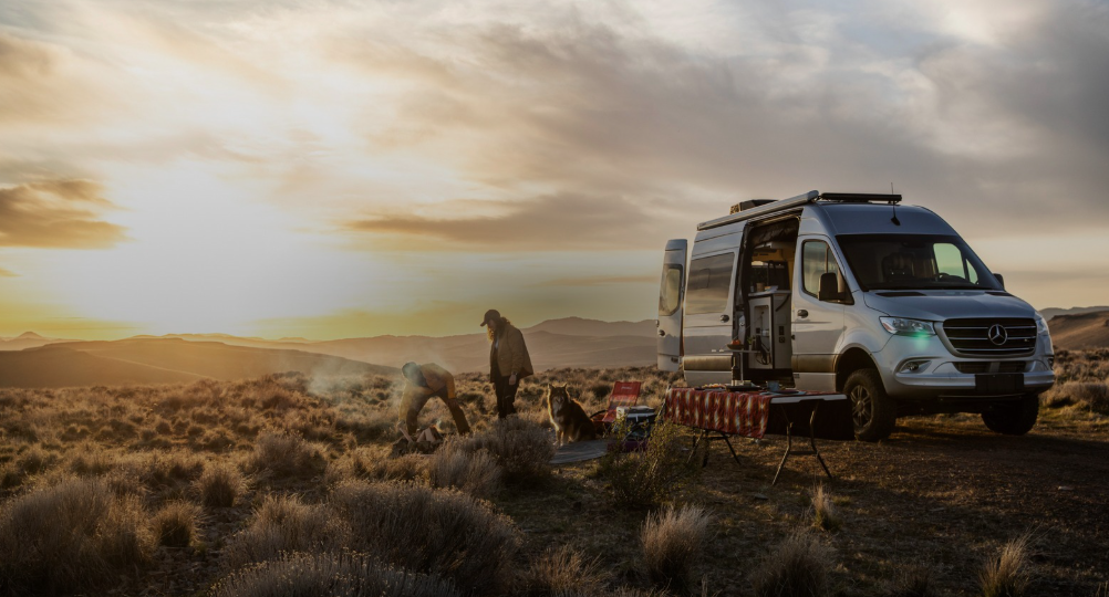 A silver Mercedes-Benz camper van is parked in a remote desert landscape during sunset. Two people tend to a small campfire near the van, while a dog sits nearby. A folding table with a red plaid tablecloth and camping chairs are set up, creating a cozy off-grid campsite scene surrounded by sagebrush and rolling hills under a glowing sky.