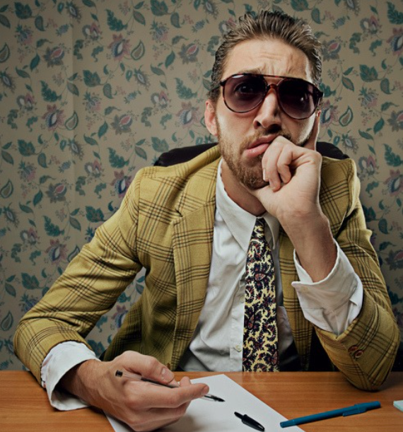 A man with a skeptical expression sits at a desk wearing large sunglasses, a yellow plaid suit jacket, and a patterned tie. He holds a pen over a blank sheet of paper with the pen cap placed nearby. The floral wallpaper and retro fashion give the scene a humorous, outdated salesman vibe.
