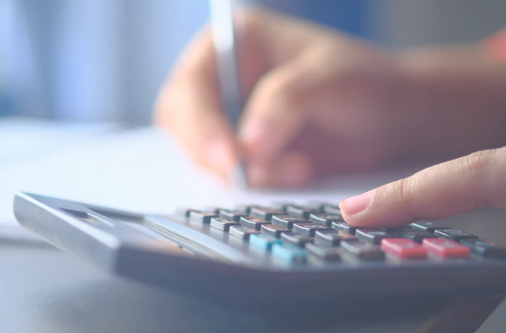 A close-up image of a person using a calculator with one hand while writing with a pen in the other. The focus is on the finger pressing the calculator buttons, with the background softly blurred. The scene suggests budgeting, financial planning, or doing calculations.