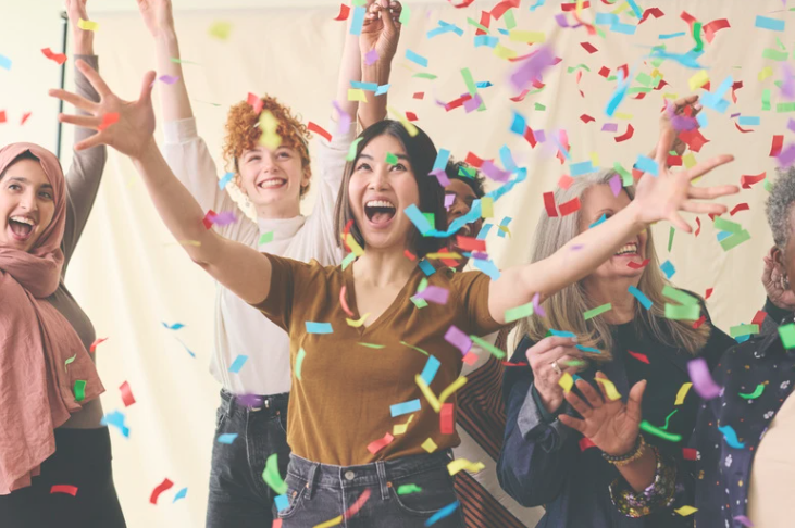 A diverse group of joyful women celebrating together indoors, surrounded by falling colorful confetti. They are smiling, laughing, and throwing their hands in the air, creating a lively and festive atmosphere. The background is neutral, keeping the focus on the energy and happiness of the group.


