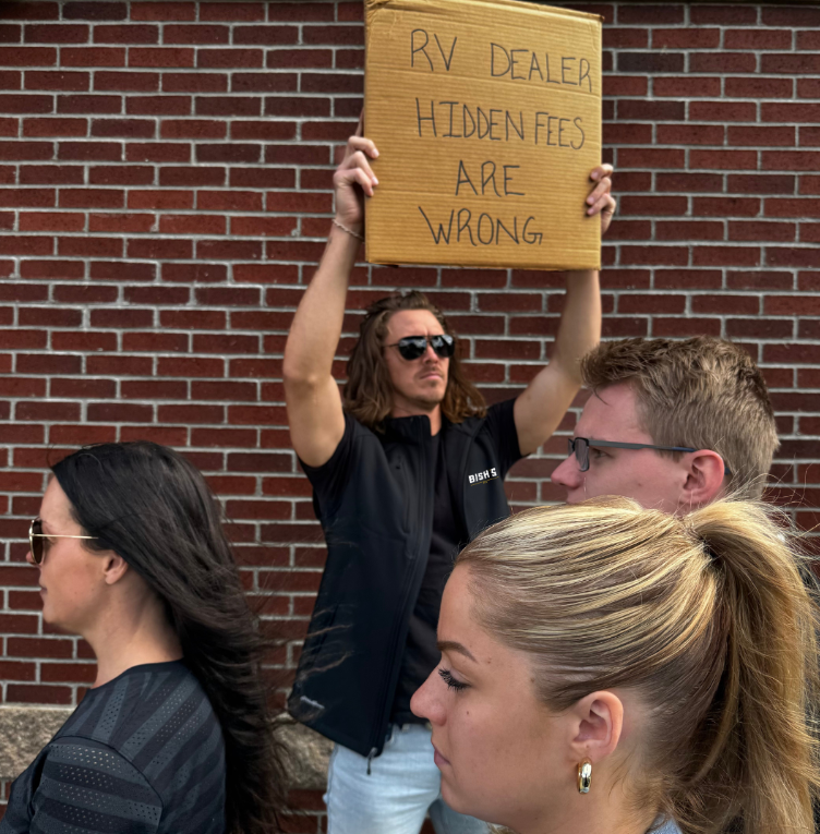 A man wearing sunglasses and a black Bish’s RV vest holds a cardboard protest sign above his head that reads, “RV DEALER HIDDEN FEES ARE WRONG.” He stands against a red brick wall while four other people walk by in the foreground, seemingly unaware. The image conveys a message about transparency in RV pricing.