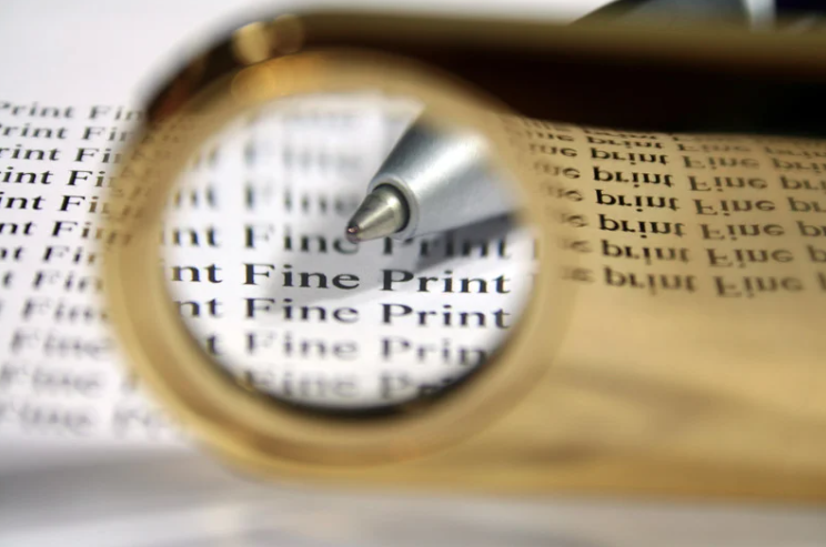 A close-up image of a magnifying glass focusing on repeated words “Fine Print” on a document. A silver pen points toward the magnified text, emphasizing the importance of reading and understanding fine print in contracts or agreements. The background is slightly blurred to draw attention to the focused area.