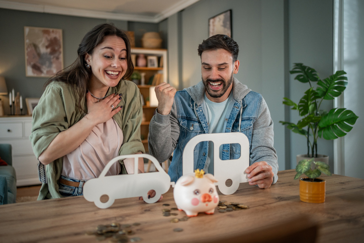 Young couple joyfully saving money in a piggy bank for their dream car and camper van, celebrating their achievement together at home and planning for a bright future