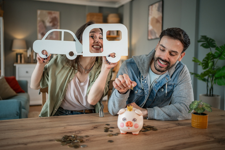 A happy couple sits at a table saving money for an RV. The man is dropping coins into a pink piggy bank with a crown, while the woman holds up playful cutouts of a car and a camper trailer. Coins are scattered across the table, and a small potted plant sits nearby. The scene suggests excitement and planning for a future RV purchase or adventure.