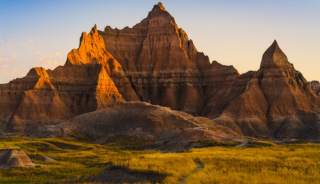 A dramatic landscape of the Badlands in South Dakota at sunset. Jagged, layered rock formations rise sharply from the grassy plains, their peaks glowing with warm golden light. The scene captures the rugged textures and unique shapes of the eroded cliffs under a clear evening sky.