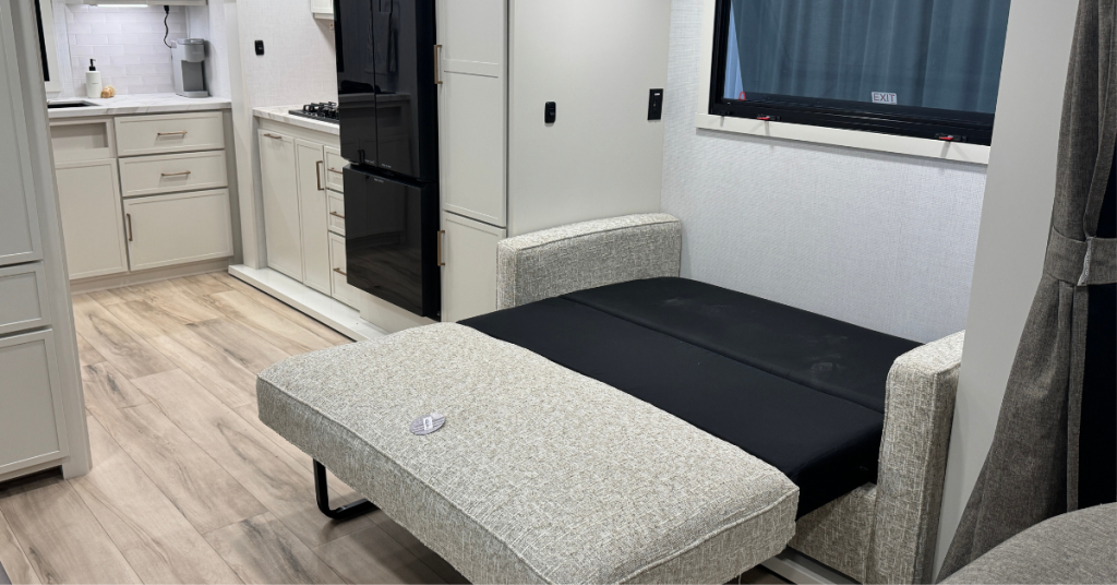 Interior view of an RV showcasing a pull-out sofa bed extended into sleeping position. The bed features a light gray textured upholstery with a black sleeping surface. In the background, the kitchen area is visible with white cabinetry, a black refrigerator, a two-burner stovetop, and light wood-look flooring throughout.