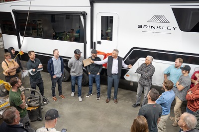 A group of people gathered in front of a white Brinkley RV during a ribbon-cutting ceremony. A man in a suit and tie is holding oversized scissors mid-cut, while others around him smile and clap. The RV has the Brinkley logo and a large window on the side.