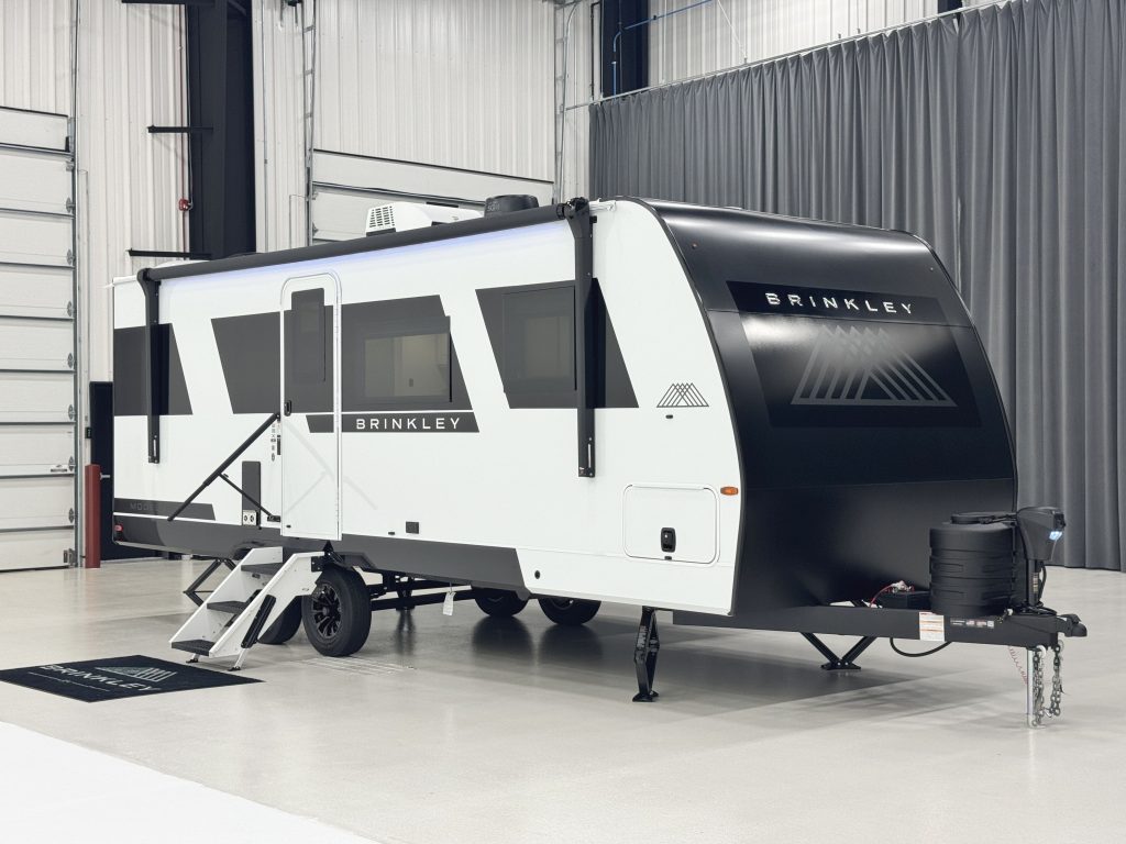 A sleek Brinkley Model Ix travel trailer is showcased indoors on a polished concrete floor. The trailer has a modern design with a white body and bold black front cap featuring the Brinkley logo. It includes large tinted windows, a side entry door with fold-out steps, dual axles, and stabilizer jacks. The setting emphasizes the trailer’s clean lines and upscale appearance.