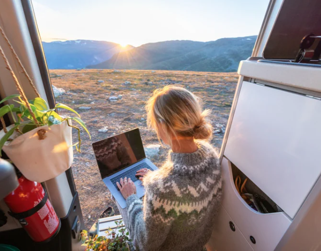 A woman sits inside an RV with the side door open, working on a laptop as the sun sets over a mountain landscape. She is wearing a gray patterned sweater, and a potted plant and fire extinguisher hang on the RV wall beside her. The scene captures a cozy remote work setup with nature views.