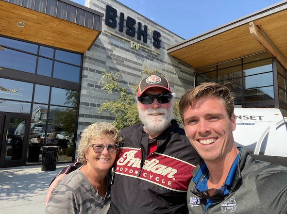 Three smiling people—two men and one woman—stand in front of a Bish’s RV dealership. The man in the center wears sunglasses, a red and black “Indian Motorcycle” jacket, and a cap. The woman on the left has curly blonde hair and glasses, and the man on the right wears a Bish’s RV shirt. Behind them is the dealership building with a large Bish’s RV sign and part of a white RV visible.