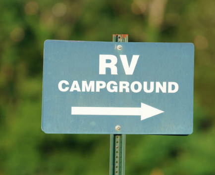 A roadside sign with a blue background and white text that reads “RV CAMPGROUND.” A large white arrow points to the right, directing travelers. The sign is mounted on a green metal pole, with blurred greenery in the background.