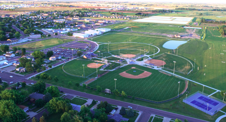 An aerial view of a large community sports complex surrounded by residential neighborhoods and open fields. The complex features multiple baseball and softball diamonds, a football/soccer field, tennis/pickleball courts, parking areas, and a small pond. Roads, houses, and other buildings are visible in the background, with farmland stretching into the distance.