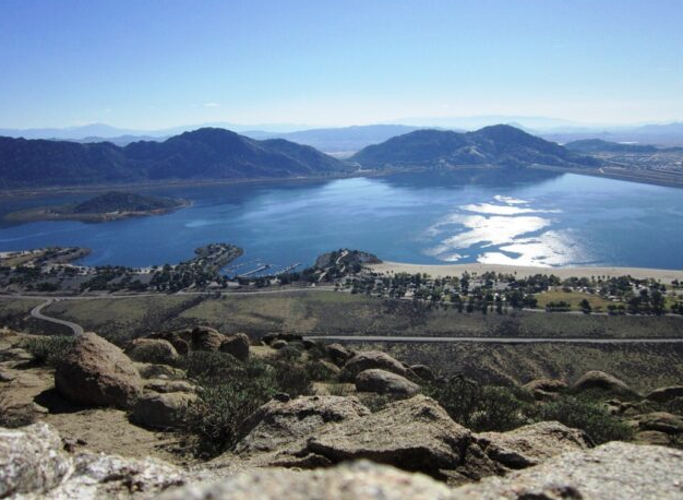 A scenic view of Lake Perris in California, surrounded by rocky hills and mountains, with campgrounds and picnic areas visible along the shoreline under a clear blue sky.