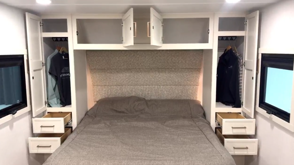 RV bedroom with a large bed covered in a gray bedspread, surrounded by white cabinetry. Open overhead storage cabinets span above the bed, while tall wardrobes with drawers are built on each side. Two windows on either side of the bed allow natural light, and recessed ceiling lights provide bright illumination. A padded, fabric-upholstered headboard adds a modern, cozy touch.