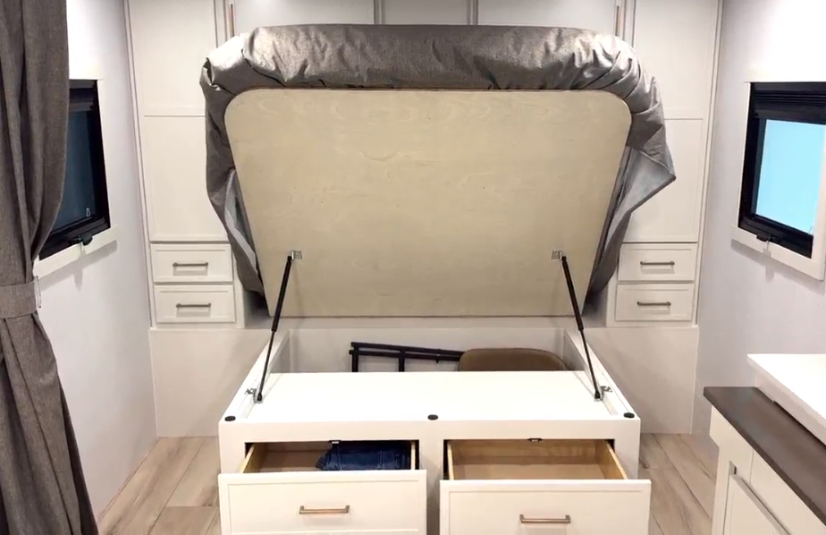 A bed in an RV is lifted to reveal under-bed storage space. The storage area includes two open drawers in the front with additional deep storage compartments underneath. The bed is supported by hydraulic lift arms, and built-in cabinetry with drawers and closets surrounds the bed on both sides.
