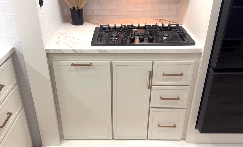 RV kitchen area with a built-in three-burner gas cooktop set into a white marble-look countertop. Above the cooktop is a stainless steel microwave, framed by light cabinetry. A small black utensil holder with wooden spoons sits in the corner against a white subway tile backsplash, with storage drawers and cabinets beneath the counter.