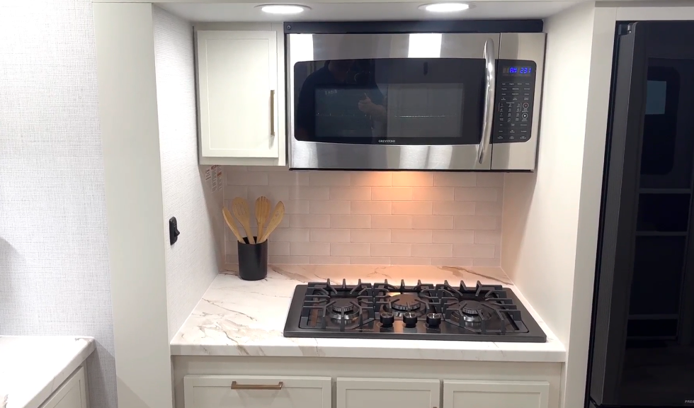 “Modern RV kitchen with a stainless steel microwave mounted above a four-burner gas cooktop, marble-look countertop, white cabinetry with brushed metal handles, and a utensil holder with wooden spoons.