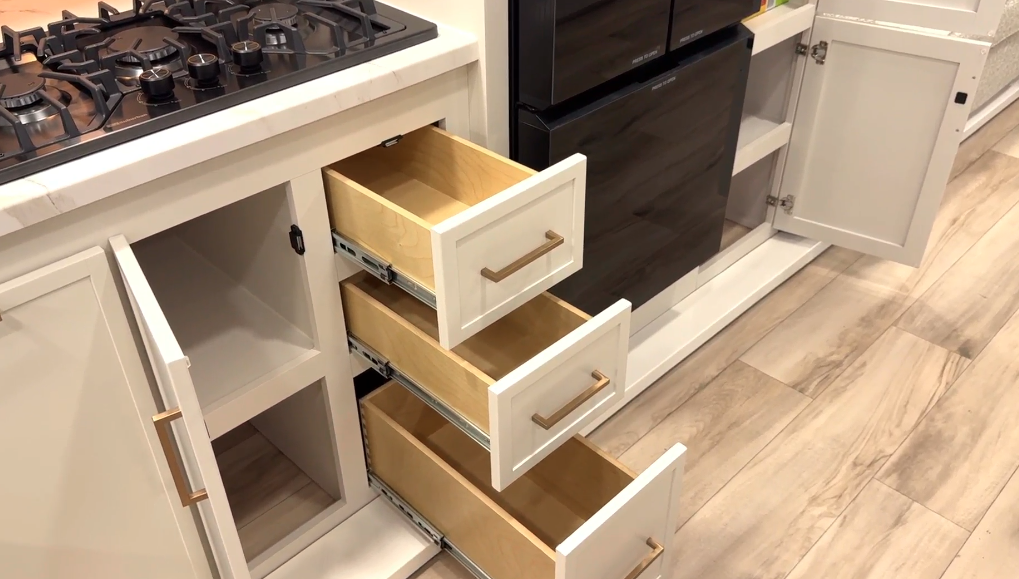 Open kitchen cabinetry and drawers inside an RV, featuring three wooden pull-out drawers with gold handles and an open lower cabinet next to a black refrigerator and stovetop above the counter. Light wood-look flooring runs throughout the space.