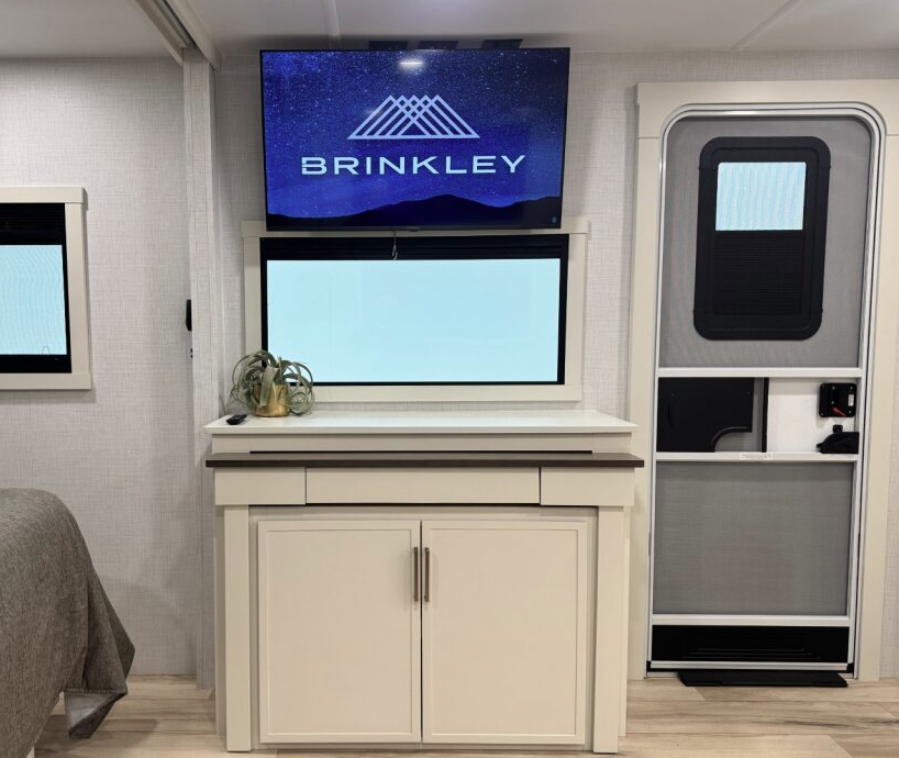 RV interior with a flat-screen TV mounted above a wide window and white cabinet storage below. A potted plant decorates the cabinet, and the entry door is positioned to the right of the entertainment center. The space has light walls and wood-look flooring.