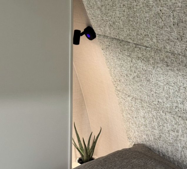 Close-up view of a Brinkley RV bedroom corner, featuring a soft gray upholstered headboard with textured fabric. A small black reading light with a purple glow is mounted on the wall above, while a green potted succulent adds a touch of decor beside the bed.