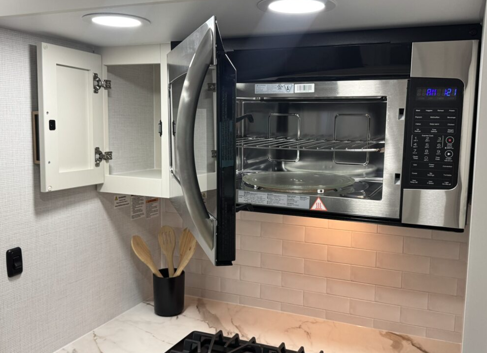 Close-up of an RV kitchen area showing a stainless steel convection microwave with its door open, revealing the interior rack and glass turntable. To the left is a small cabinet with its door open, and below sits a marble-look countertop with a black utensil holder containing wooden spoons. The backsplash features light subway tiles.”