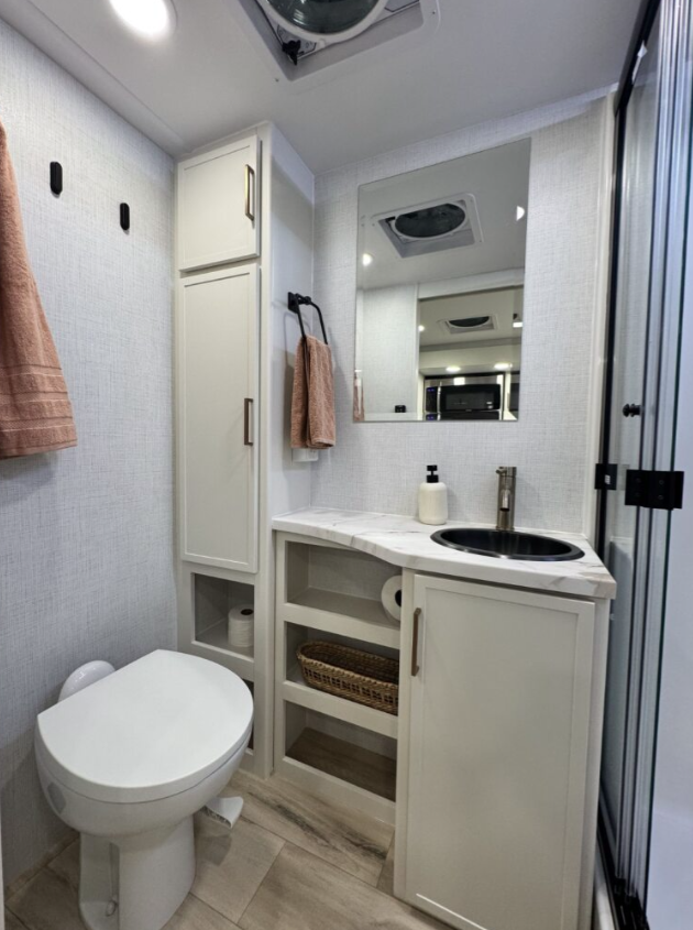 Compact RV bathroom featuring a modern design with a toilet, built-in storage cabinet, open shelving with a wicker basket and toilet paper, a marble-look countertop with an under-mounted round sink, a faucet, and a wall-mounted mirror. Neutral tones, a towel rack with a hanging peach-colored towel, and a glass shower door create a clean, functional layout.