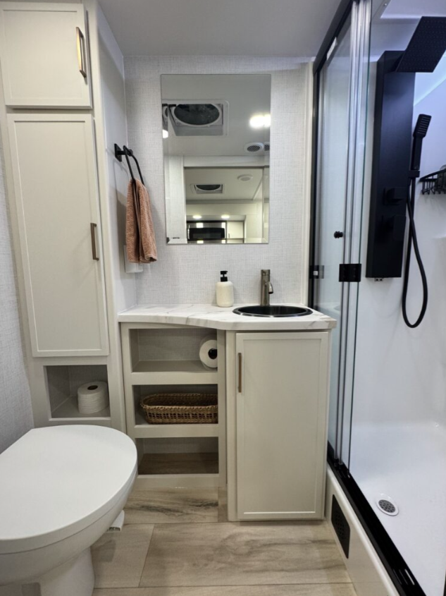 Modern RV bathroom with a toilet, vanity sink, and mirrored medicine cabinet. The vanity features open shelving with storage baskets and a toilet paper holder, while the corner shower with a glass door and black fixtures is visible on the right.