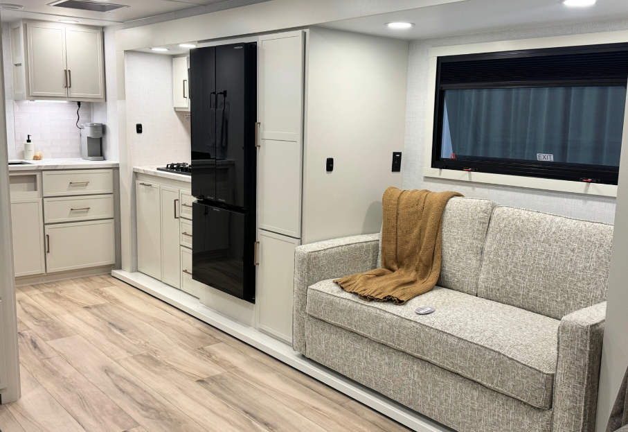 Modern RV interior featuring a light gray sofa with a brown throw blanket, a black residential-style refrigerator, and a full kitchen with white cabinetry, drawers, and a stainless-steel sink. The space has wood-look flooring and large windows for natural light.