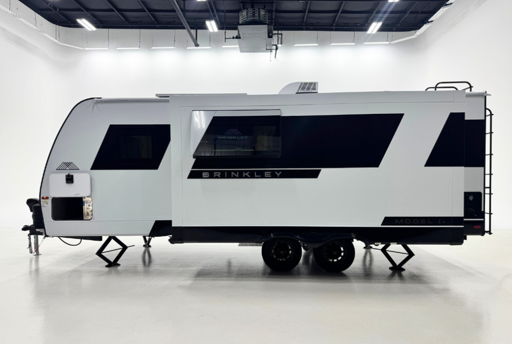 Side view of a modern Brinkley Model 1x travel trailer in a bright studio setting. The RV features a sleek white exterior with bold black geometric accents, tinted windows, dual axles, and stabilizer jacks. A rear ladder is visible on the right side.