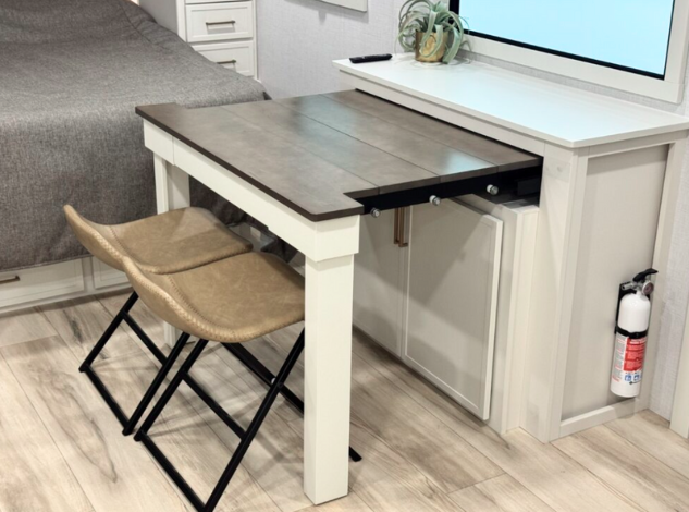 A compact RV dining setup featuring a small rectangular table with a wood-toned surface and white legs. Two tan cushioned stools with black metal frames are tucked underneath. The table is positioned in front of a white storage cabinet with a potted plant on top, next to a large window. A bed is partially visible in the background, and a fire extinguisher is mounted to the side of the cabinet.