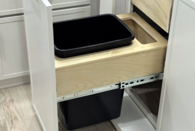 Pull-out kitchen cabinet with a dual-bin waste system inside an RV. The drawer features a wooden frame holding a removable black bin on top, with a second larger bin positioned below for trash or recycling.