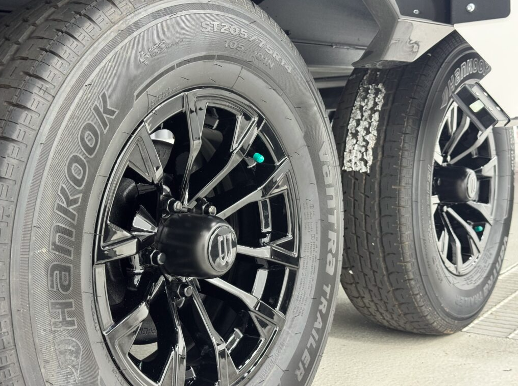 Close-up view of two Hankook Vantra Trailer tires with glossy black alloy rims, mounted on a tandem axle of an RV. The tires feature visible tread detail, green valve stem caps, and part of the RV’s undercarriage above.