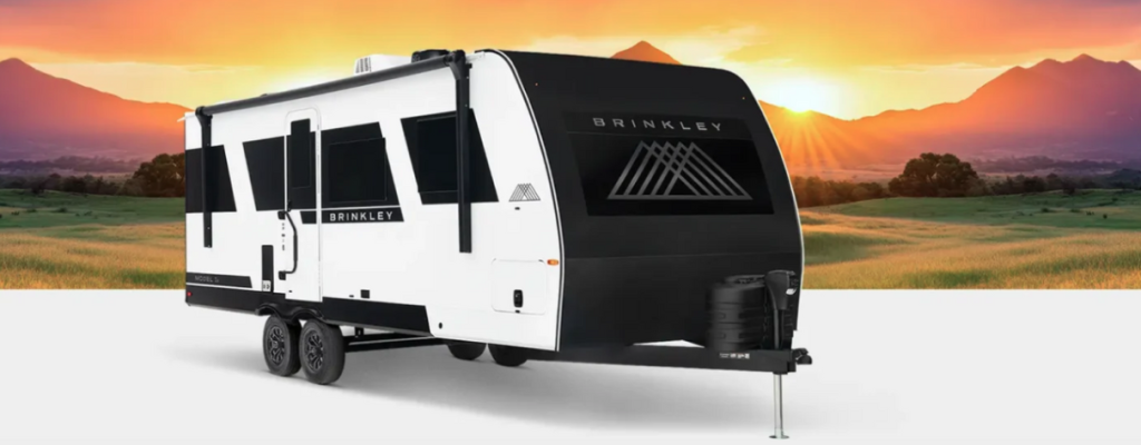 A sleek Brinkley Model Ix travel trailer is parked in front of a scenic mountain landscape at sunrise. The trailer features a modern white and black exterior design with bold geometric graphics, dual axles, and propane tanks mounted at the front.