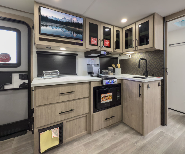 Modern RV kitchen interior featuring light wood cabinetry with black hardware, a white solid-surface countertop, and a black faucet. The kitchen is equipped with a built-in microwave, stovetop with glass cover, and stainless steel range hood. Overhead cabinets with glass inserts provide storage, while under-counter drawers add functionality. A flat-screen TV is mounted above the counter, and the space is accented by modern lighting and a dark textured backsplash. The design combines functionality with a contemporary home-like feel.